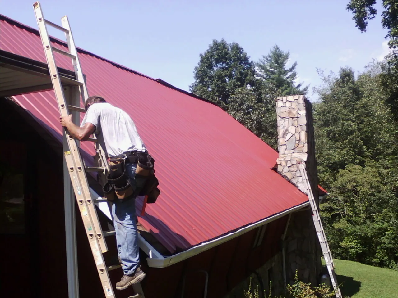Roofer on a ladder beside a red metal roof and stone chimney — Soffit & Fascia Repair by Roofing Brothers in South Mills